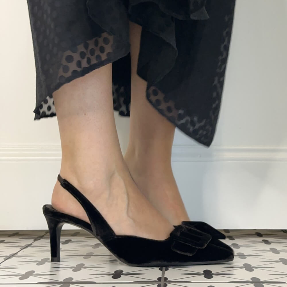 Lodi Testa Brown velvet slingback kitten heel pumps with bow details, shown on a model wearing black patterned dress against a white wall and tiled floor.