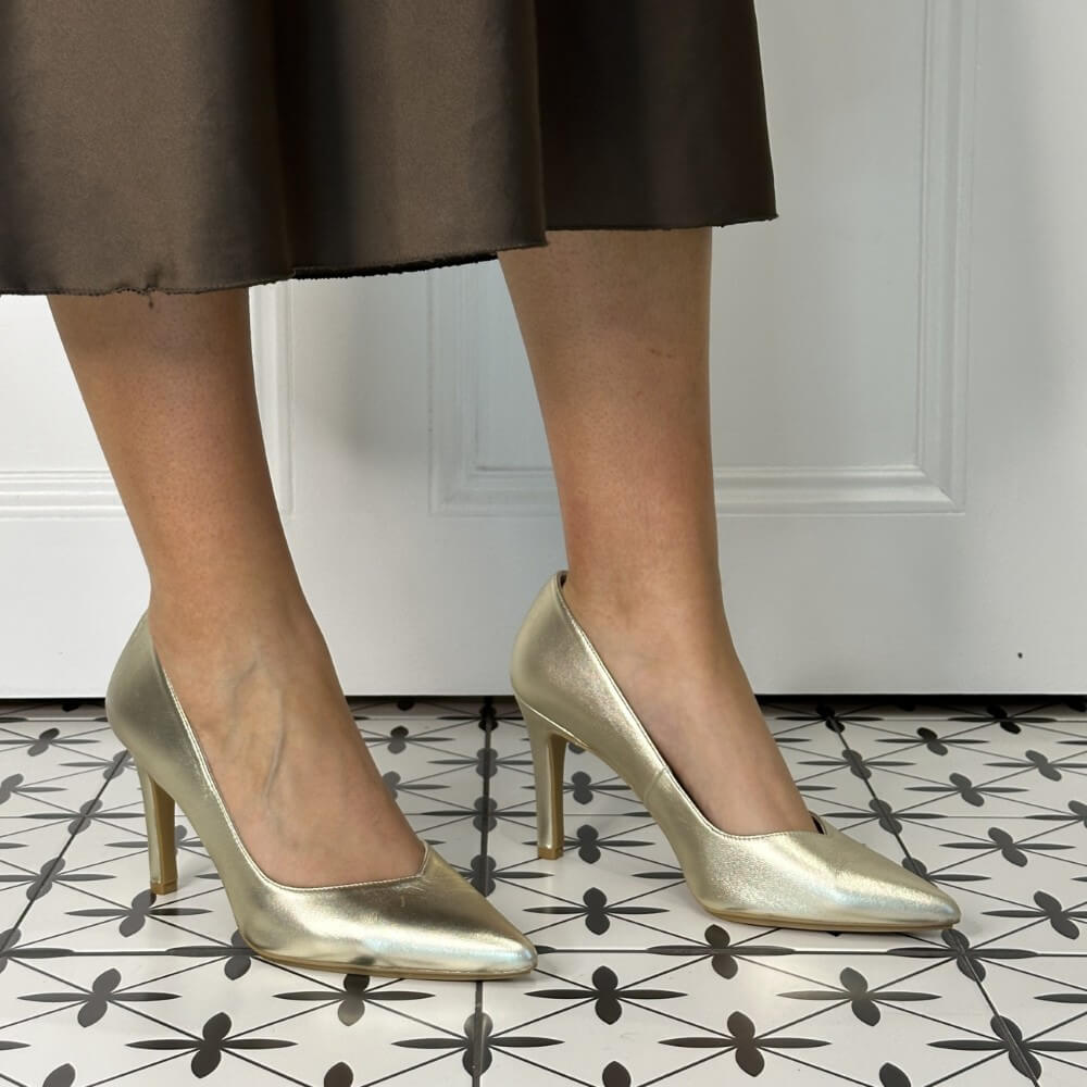 Gold high-heeled shoes worn with a brown skirt on a patterned floor.
