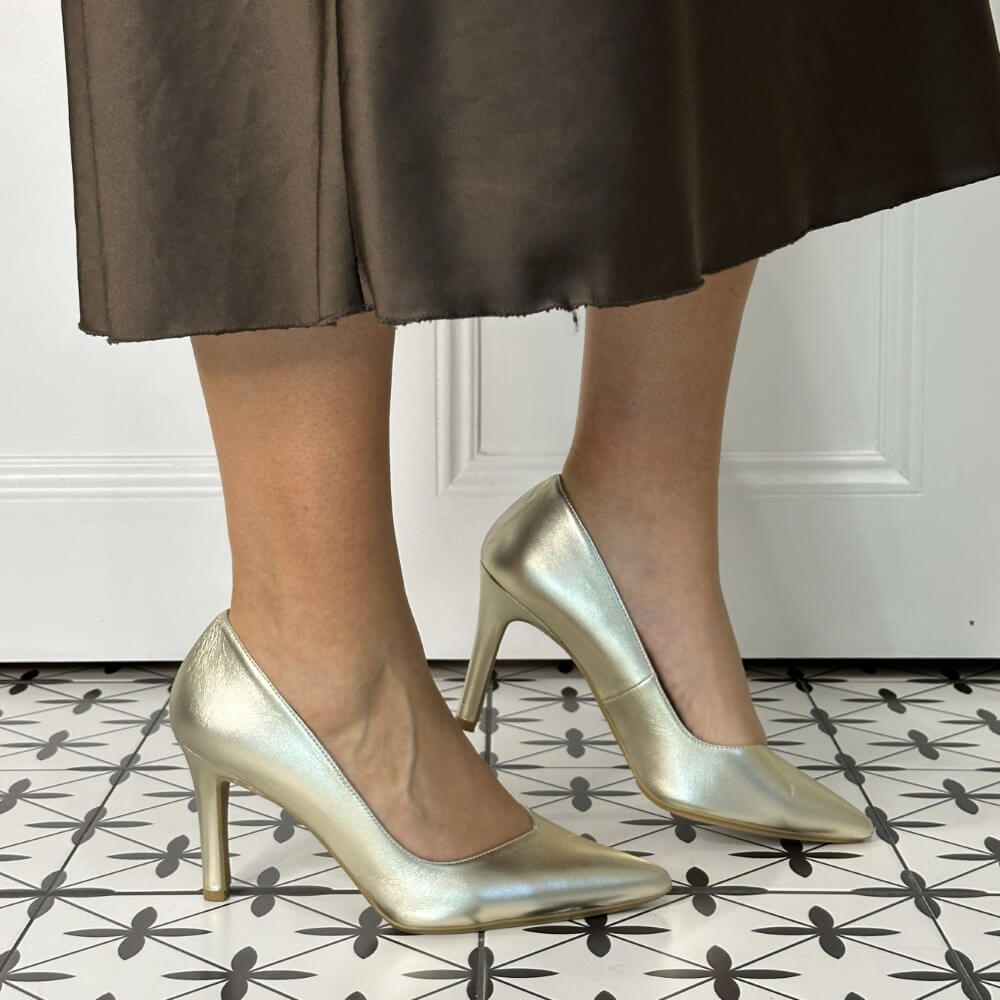 Gold high-heeled shoes worn with a brown skirt on a patterned floor.