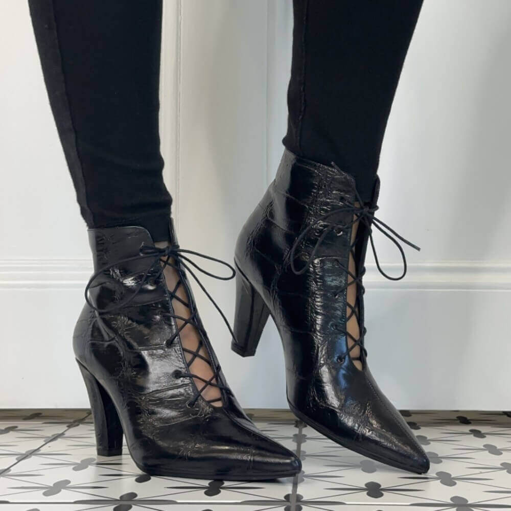 Lodi Black high-heeled ankle boots with a glossy crocodile-embossed texture and lace-up front, shown on a model wearing black leggings against a white wall and patterned tile floor.