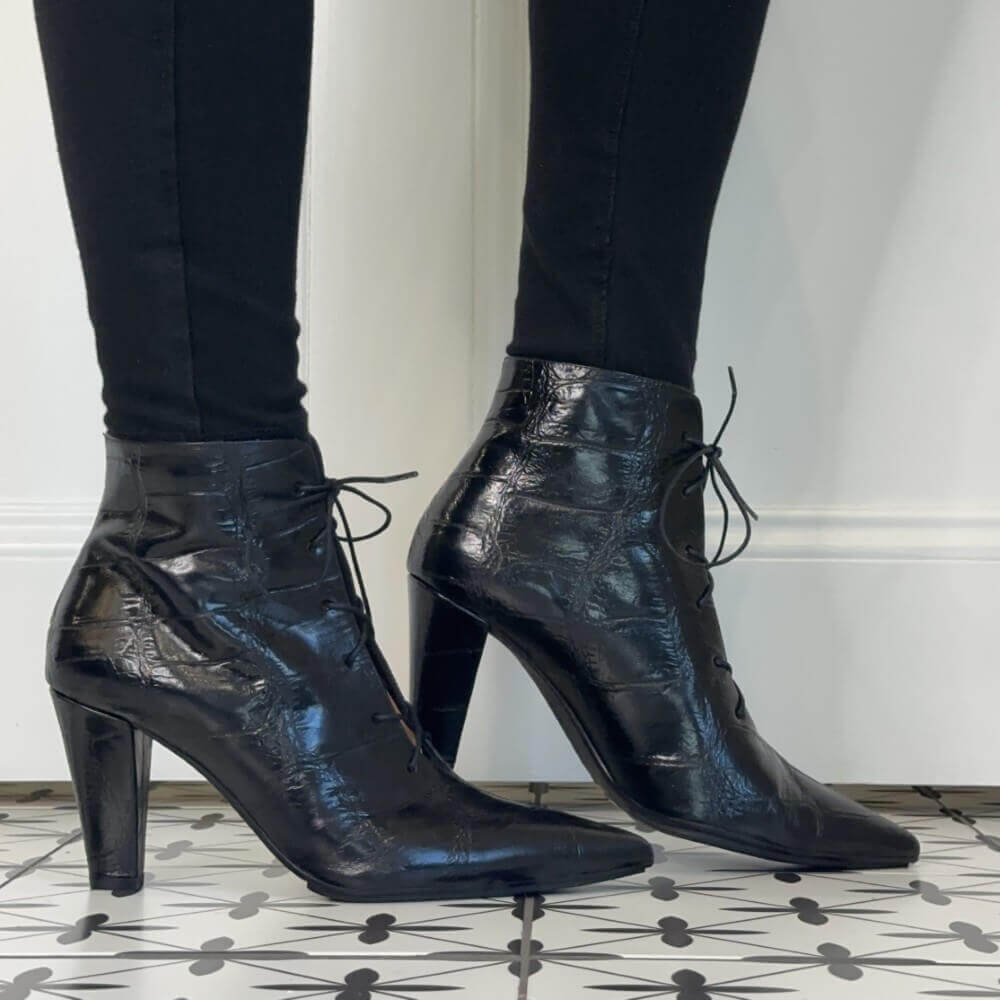 Lodi Black high-heeled ankle boots with a glossy crocodile-embossed texture and lace-up front, shown on a model wearing black leggings against a white wall and patterned tile floor.