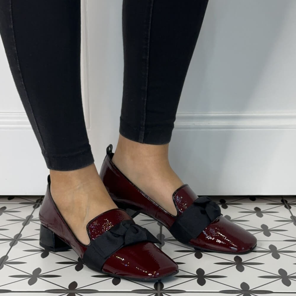 Burgundy shoes with black bows worn with black pants on a patterned floor.