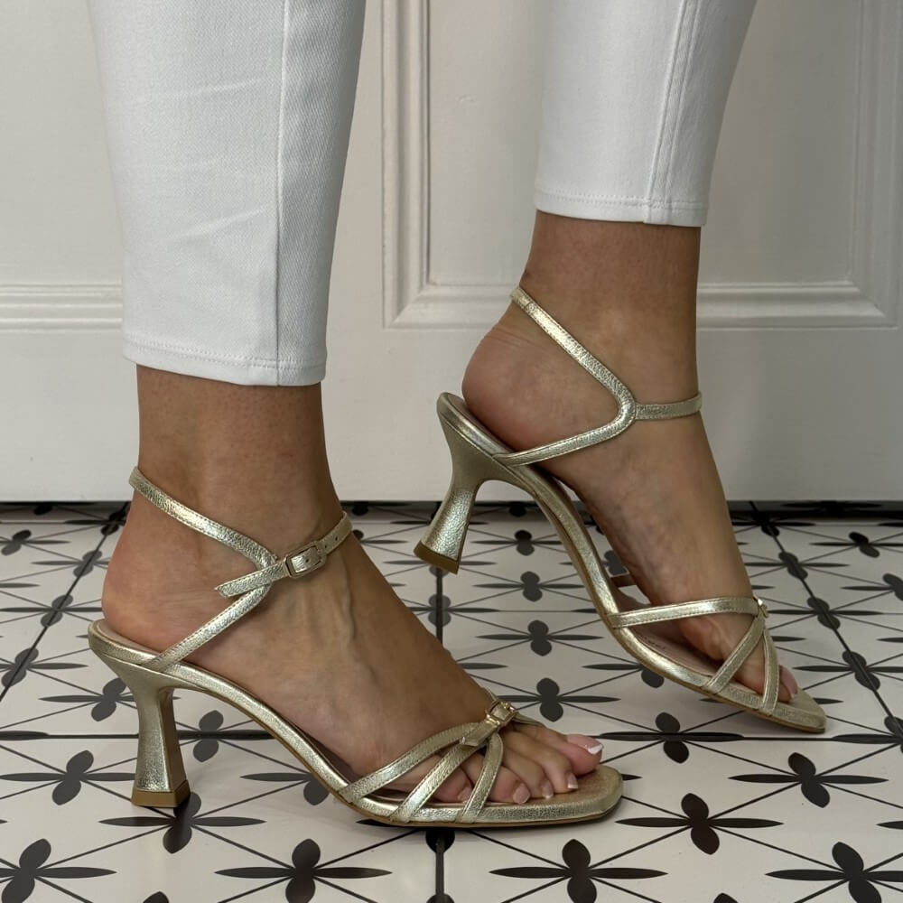 Lodi Gold high-heeled sandals worn with white pants on a patterned floor.