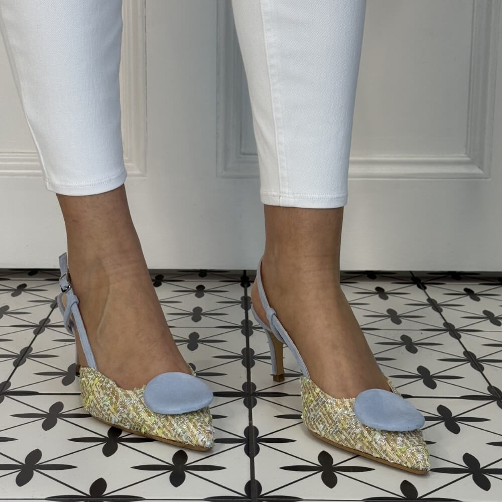 High-heeled shoes with glittery design and light blue accents worn with white pants on a patterned floor.