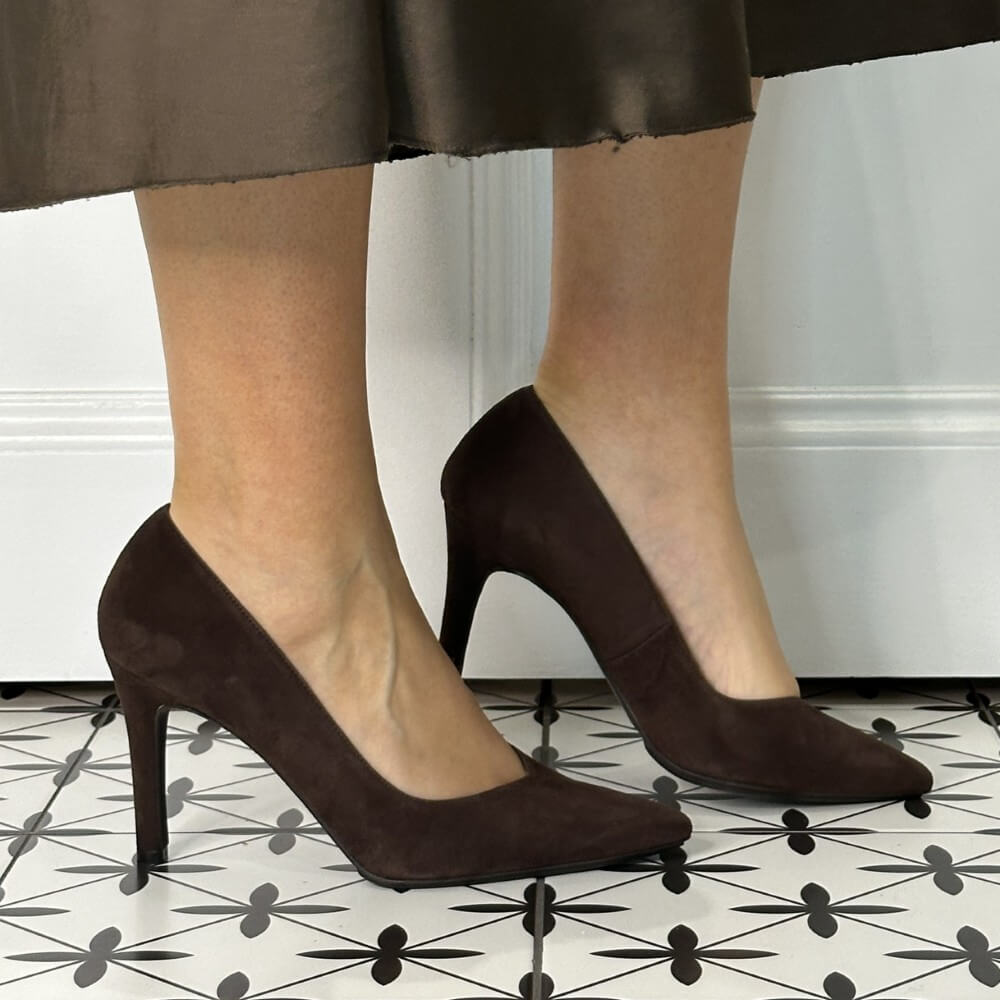 Brown high-heeled shoes worn with a skirt on a patterned floor.