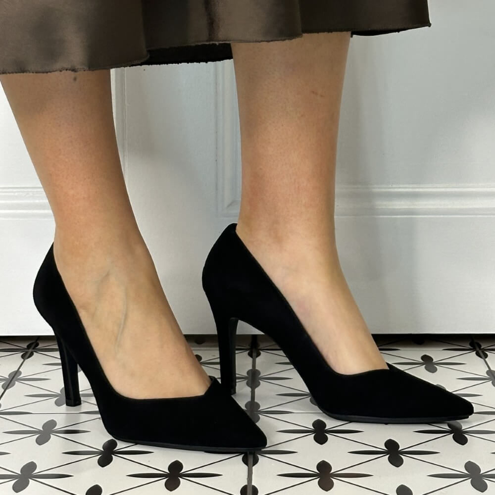 Black high-heeled shoes worn with a brown skirt on a patterned floor.