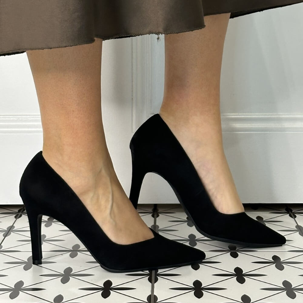 Black high-heeled shoes worn with a brown skirt on a patterned floor.