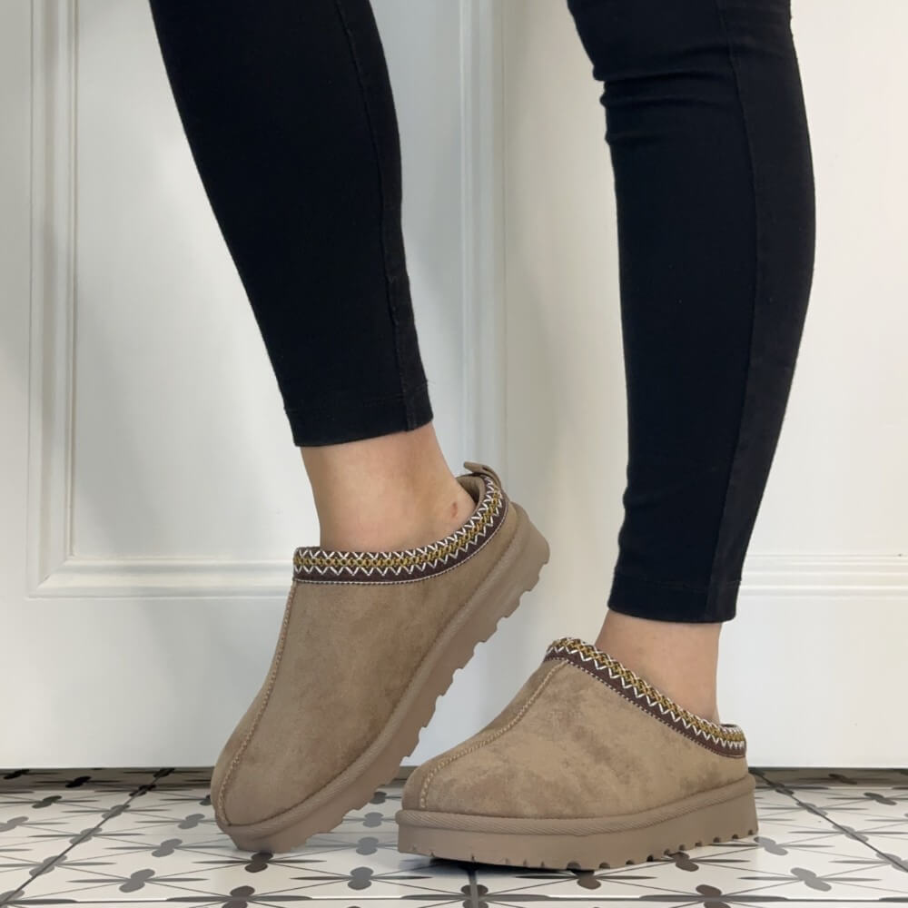 A person wearing black leggings and Lunar taupe suede slip-on slippers with a braided trim detail, standing on a patterned tile floor in front of a white paneled wall.
