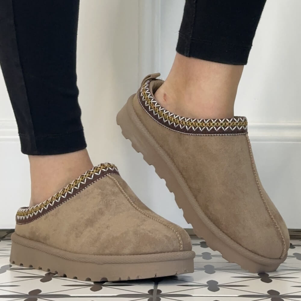 A person wearing black leggings and Lunar taupe suede slip-on slippers with a braided trim detail, standing on a patterned tile floor in front of a white paneled wall.