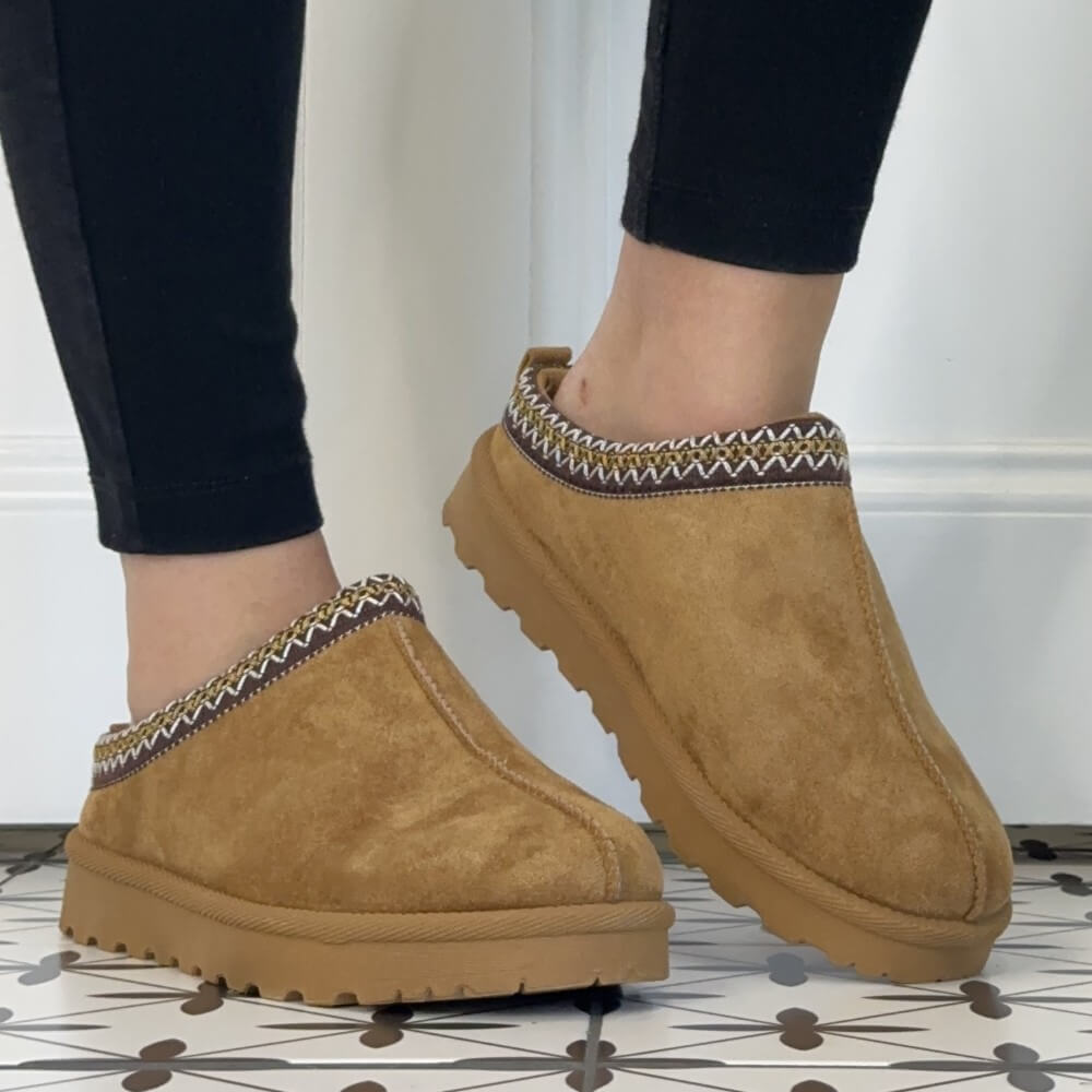 A pair of Lunar tan suede slip-on clog  slippers with a patterned fabric trim, shown being worn with black leggings on a patterned tile floor.