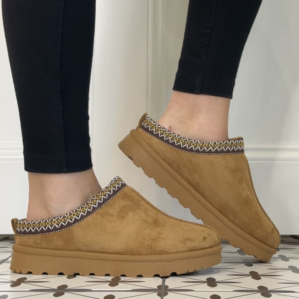 A pair of Lunar tan suede slip-on clog  slippers with a patterned fabric trim, shown being worn with black leggings on a patterned tile floor.