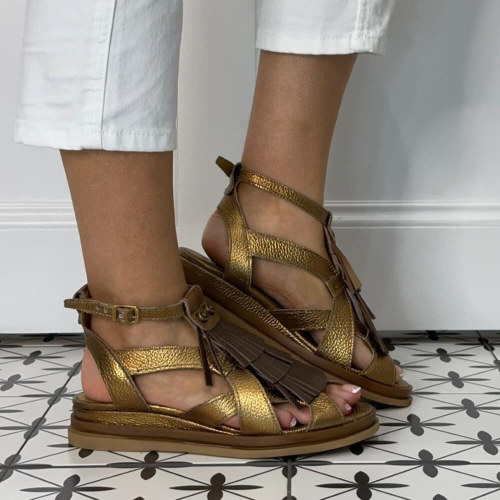 MJUS Gladiator Bronze Gold sandals with brown fringes worn by a person on a patterned floor.