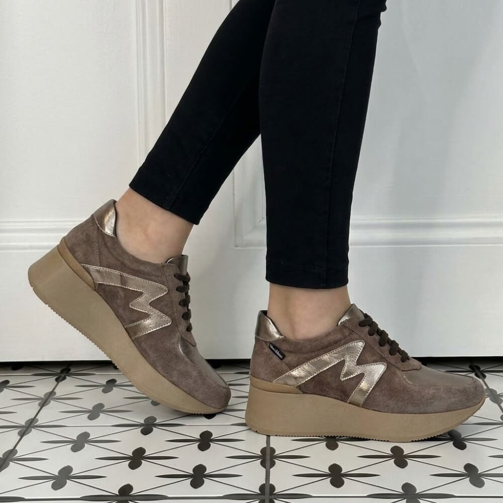 A person wearing Marco Moreo brown suede platform sneakers with metallic silver 'M' logos, paired with black pants, standing on a black and white patterned tile floor against a white panelled wall.