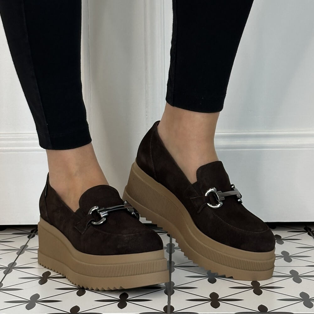 A person wearing Marco Moreo dark brown platform slip-on shoes with a small silver horse bit detail on the strap, paired with black leggings, standing on a black and white patterned tile floor.