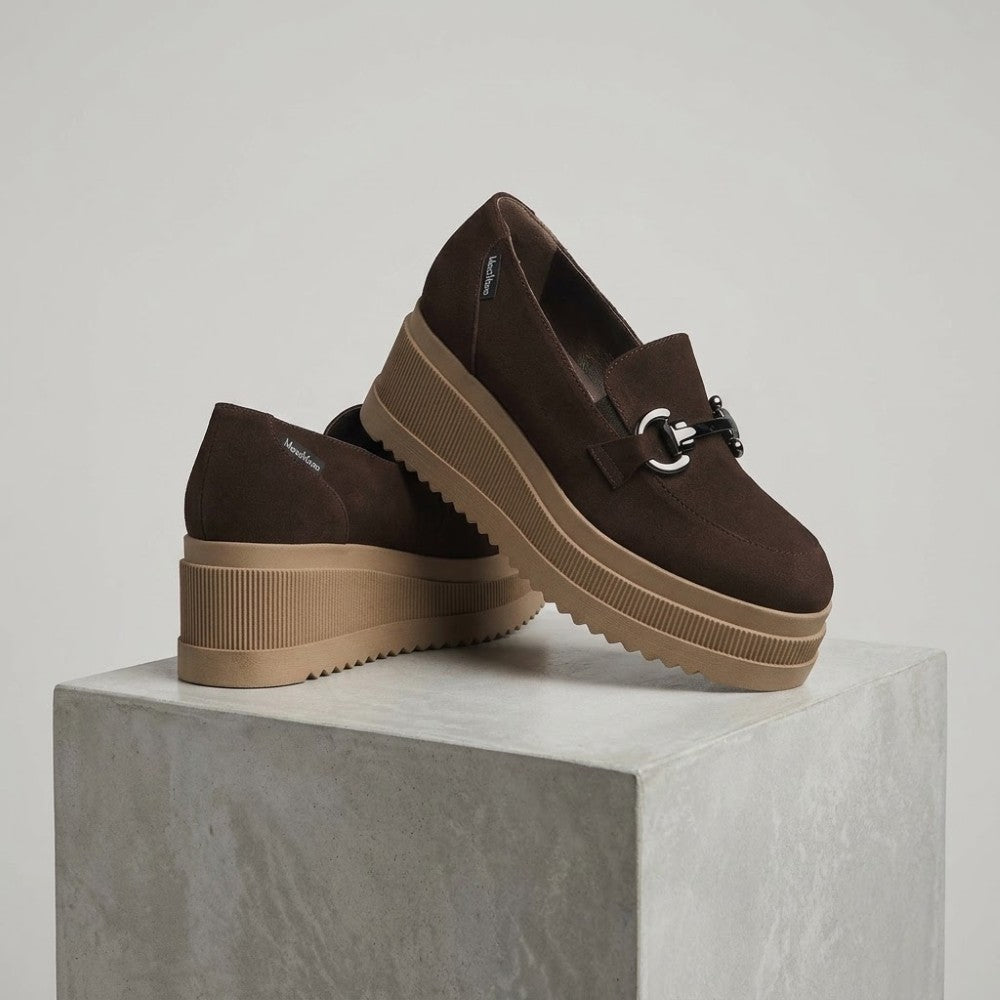 A pair of dark brown platform slip-on shoes with a chunky tan sole and a silver-tone horsebit detail on the toe, displayed on a light gray concrete block against a neutral studio background.