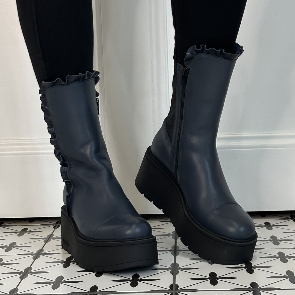 A pair of Marco Moreo dark navy blue platform ankle boots with ruffled trim and side zippers, worn with black leggings on a patterned black and white tile floor.