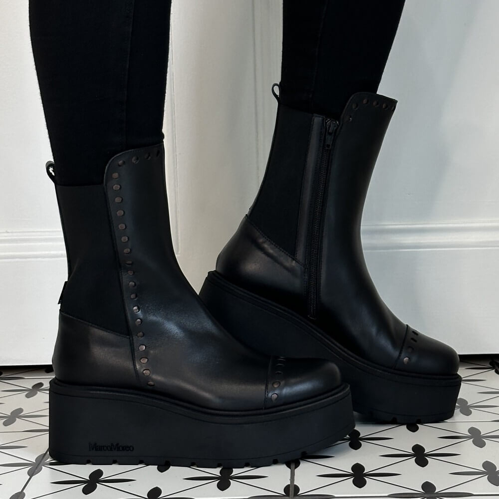 Marco Moreo Black platform ankle boots with elastic side panels and decorative Punched dots, shown worn with black leggings on a patterned tile floor against a white wall.