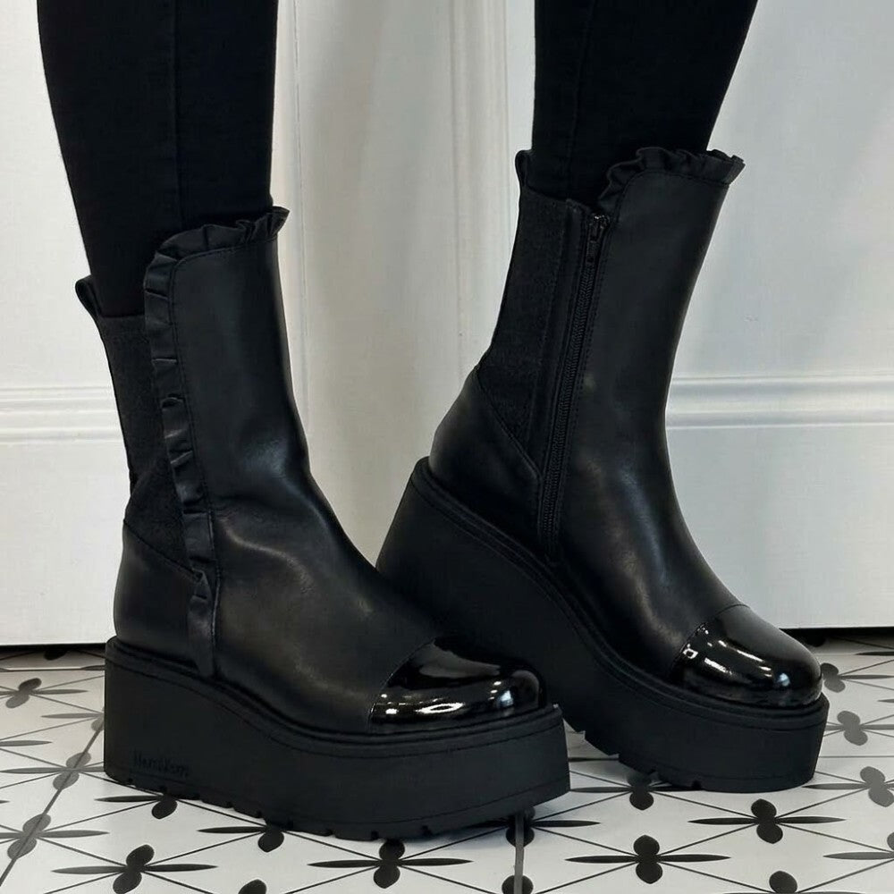Marco Moreo Black platform ankle boots with glossy patent toe caps, ruffled trim, and side zippers, shown on a model wearing black leggings on a black-and-white patterned tile floor.