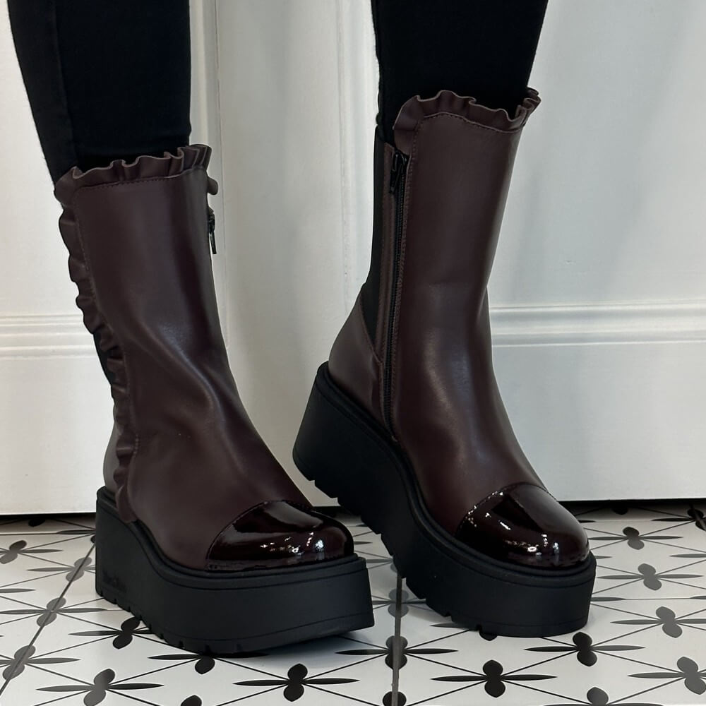 A pair of Marco Moreo dark wine ankle boots with black platform soles, ruffled trim, and side zippers, shown on a model wearing black leggings on a black and white patterned tile floor.