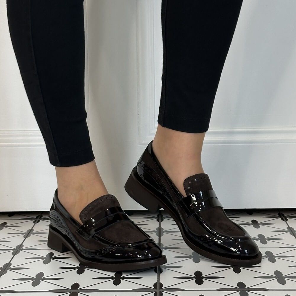 A person wearing Marco Moreo brown patent leather and suede penny loafers with low block heels, paired with black skinny jeans, standing on a black and white patterned tile floor in front of a white panelled wall.