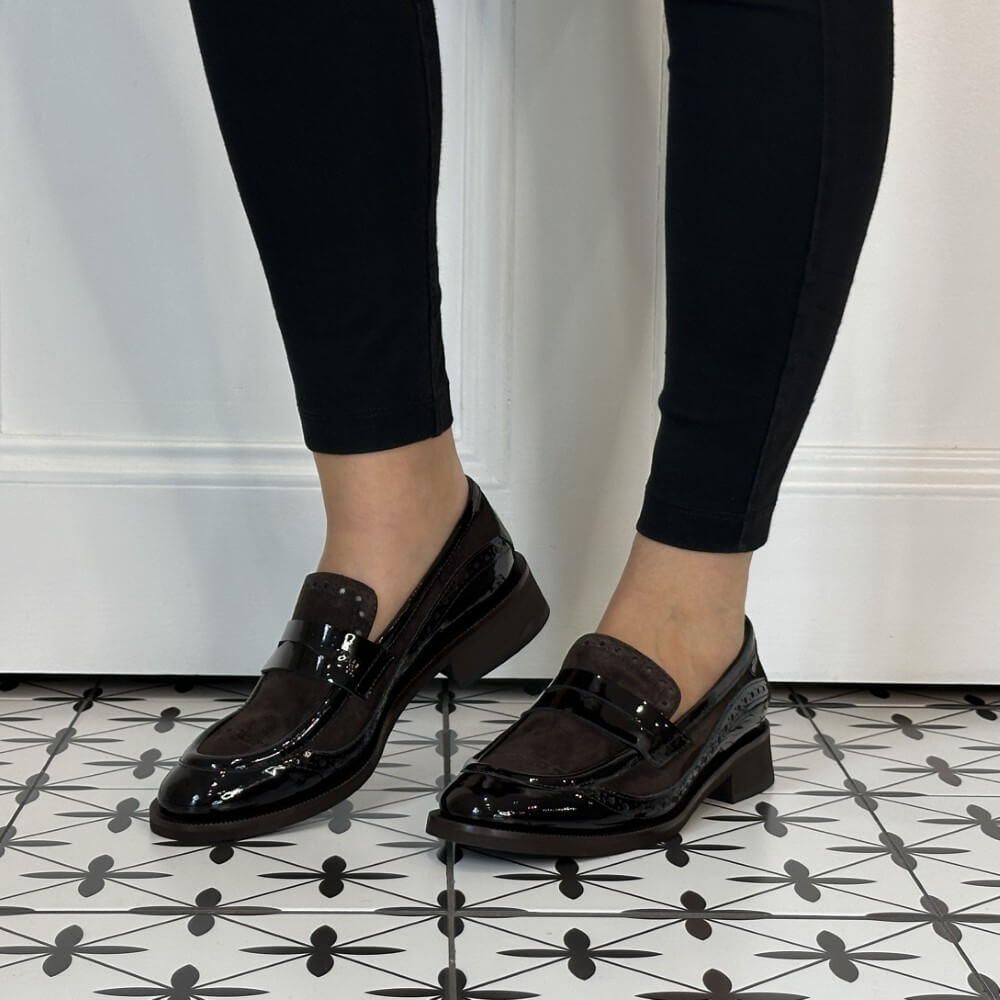 A person wearing Marco Moreo brown patent leather and suede penny loafers with low block heels, paired with black skinny jeans, standing on a black and white patterned tile floor in front of a white panelled wall.