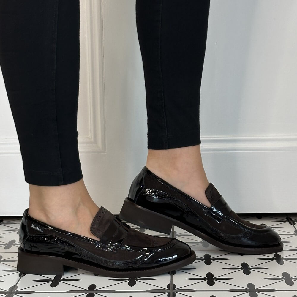 A person wearing Marco Moreo brown patent leather and suede penny loafers with low block heels, paired with black skinny jeans, standing on a black and white patterned tile floor in front of a white panelled wall.