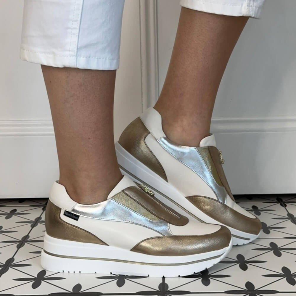 Marco Moreo White and gold sneakers worn by a person on a patterned floor.