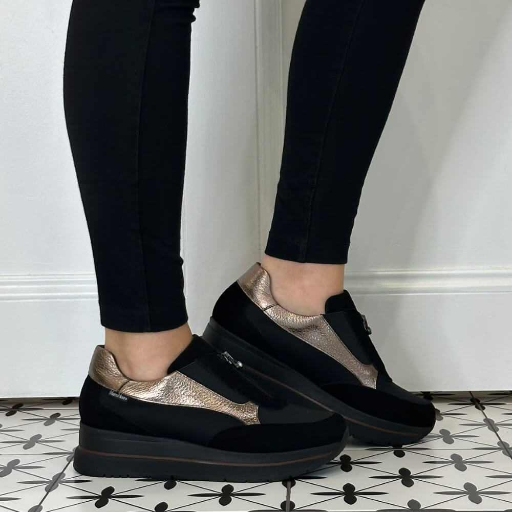 A person wearing Marco Moreo black slip-on platform sneakers with metallic rose gold panels, paired with black leggings, standing on a black and white patterned tile floor against a white wall.