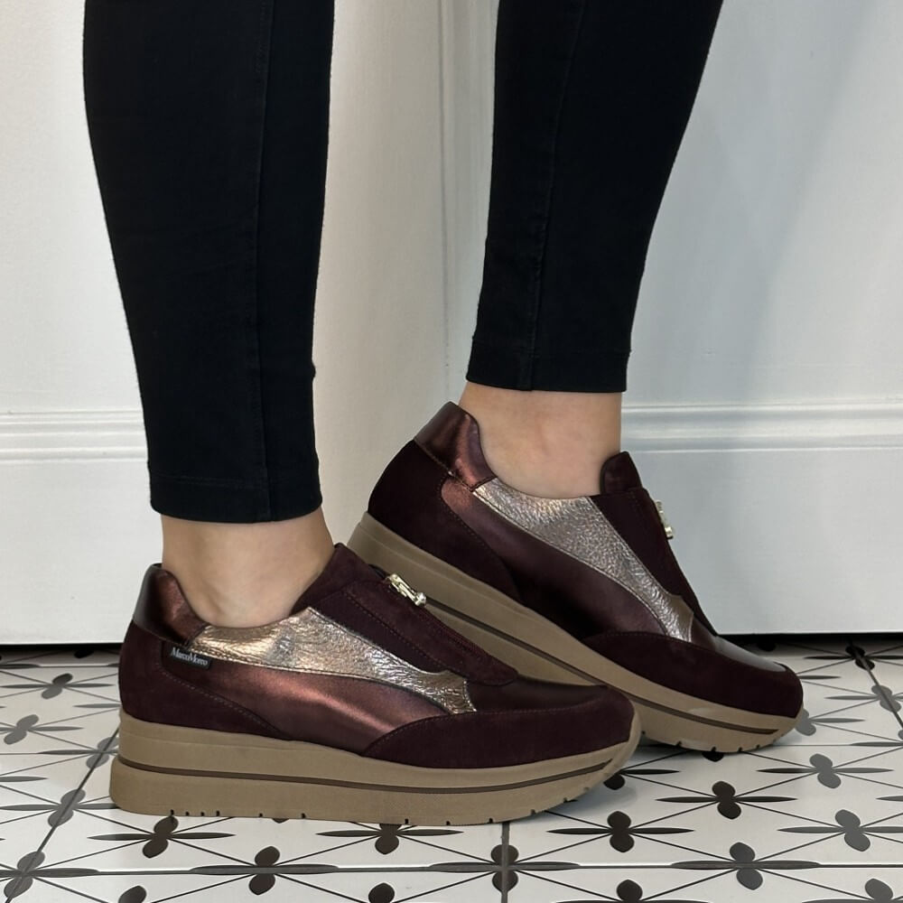 A person wearing Marco Moreo dark burgundy slip-on platform sneakers with metallic silver panels and black leggings, standing on a black-and-white patterned tile floor.