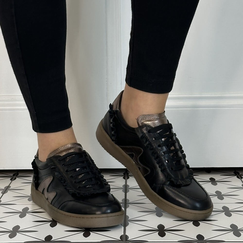 A pair of Marco Moreo black leather sneakers with metallic rose gold accents and black ruffle trim, shown on a model wearing black leggings on a patterned tile floor.
