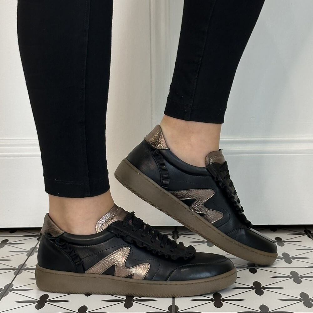 A pair of Marco Moreo black leather sneakers with metallic rose gold accents and black ruffle trim, shown on a model wearing black leggings on a patterned tile floor.