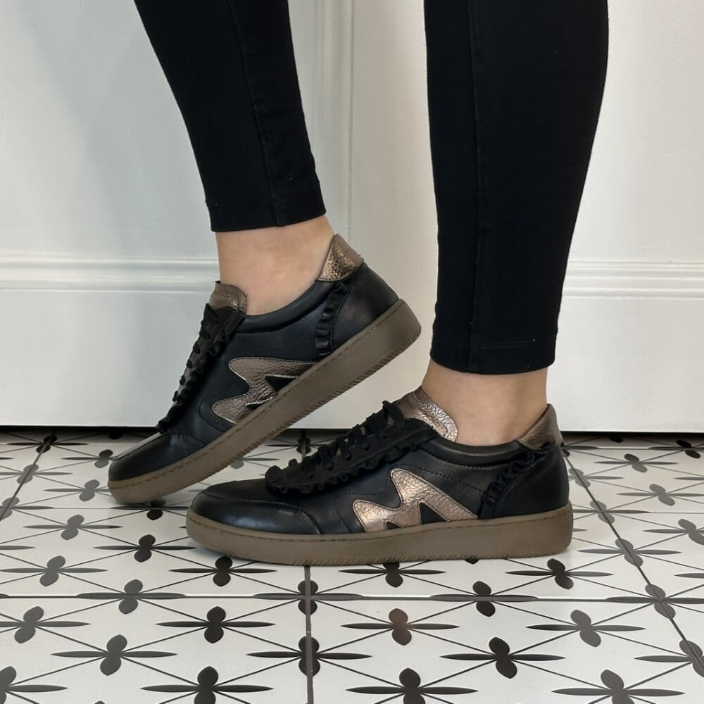A pair of Marco Moreo black leather sneakers with metallic rose gold accents and black ruffle trim, shown on a model wearing black leggings on a patterned tile floor.