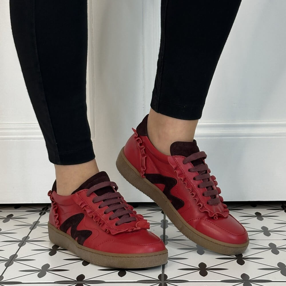 A person wearing Marco Moreo red leather sneakers with dark brown details, brown laces, and brown soles, paired with black leggings, standing on a black and white patterned tile floor.