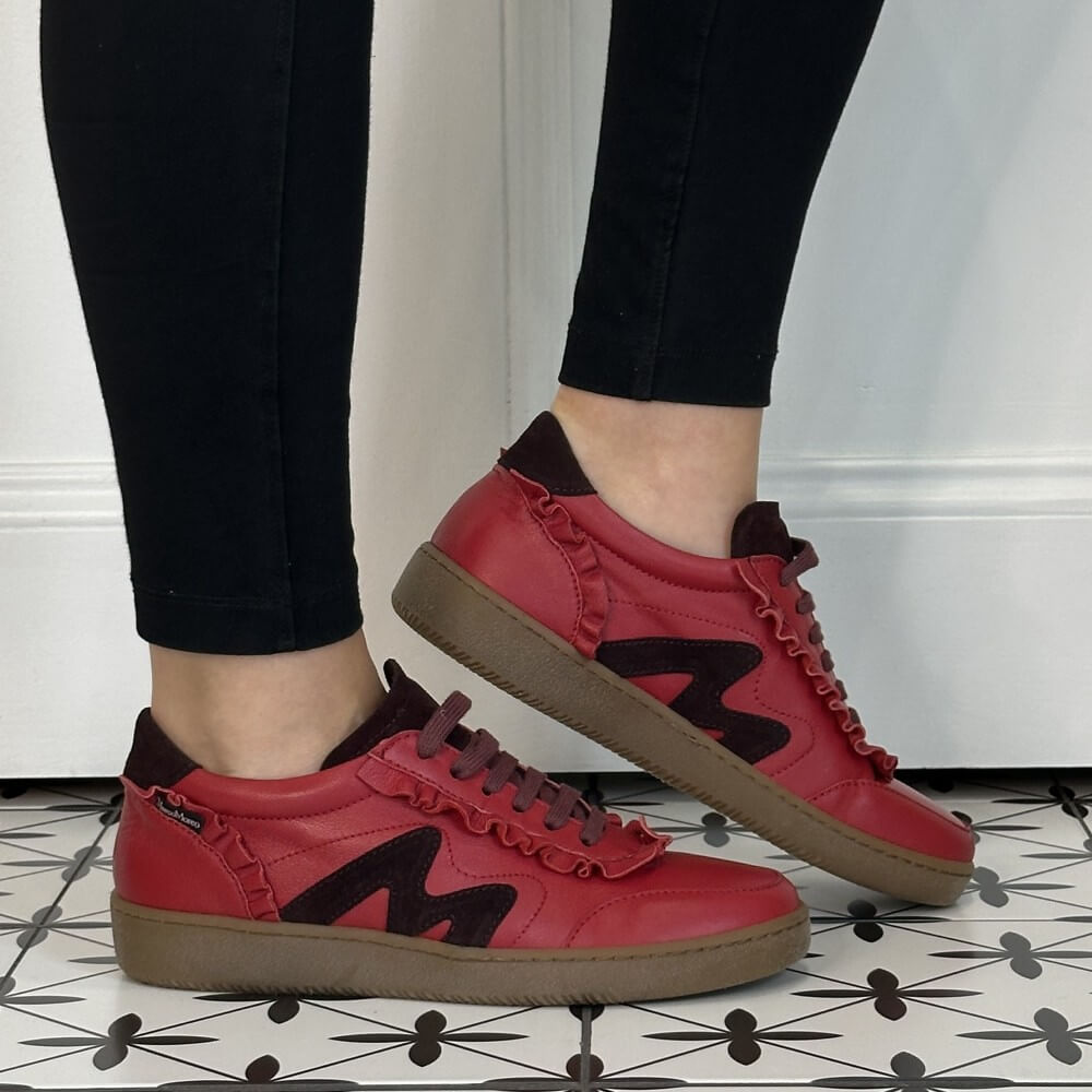 A person wearing Marco Moreo red leather sneakers with dark brown details, brown laces, and brown soles, paired with black leggings, standing on a black and white patterned tile floor.