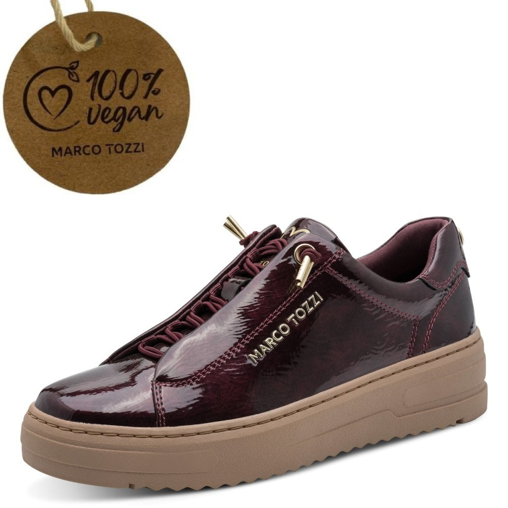 A low-top platform vegan sneaker in glossy dark burgundy patent  with matching burgundy laces, a tan sole, and gold-tone hardware. A round brown paper tag reading '100% vegan MARCO TOZZI' is shown in the upper left on a white background.