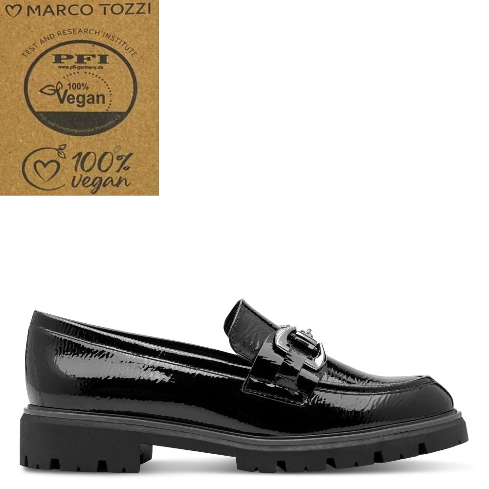 A black patent vegan leather loafer by Marco Tozzi, featuring a decorative metal buckle strap and a chunky lug sole, shown against a white background with a brown '100% Vegan' certification badge in the top-left corner.
