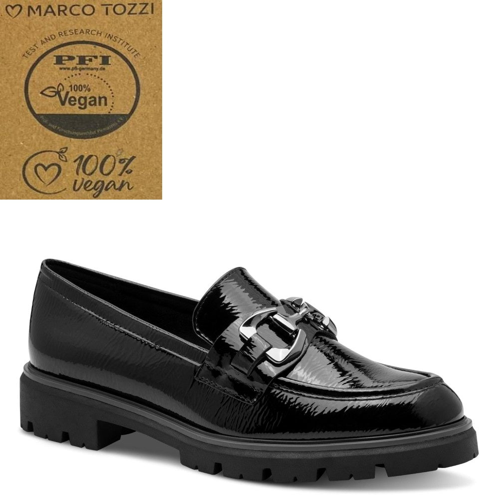 A black patent vegan leather loafer by Marco Tozzi, featuring a decorative metal buckle strap and a chunky lug sole, shown against a white background with a brown '100% Vegan' certification badge in the top-left corner.