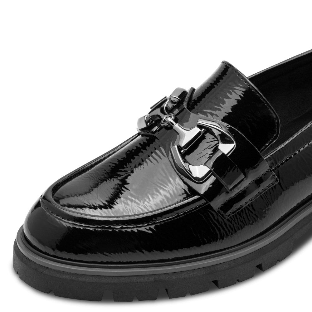 A black patent vegan leather loafer by Marco Tozzi, featuring a decorative metal buckle strap and a chunky lug sole, shown against a white background 