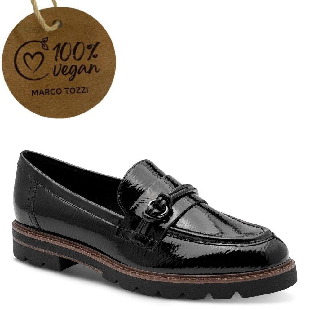 A black patent leather loafer shoe by Marco Tozzi, shown from a three-quarters angle on a white background, with a brown paper tag in the upper left corner reading '100% vegan MARCO TOZZI'.