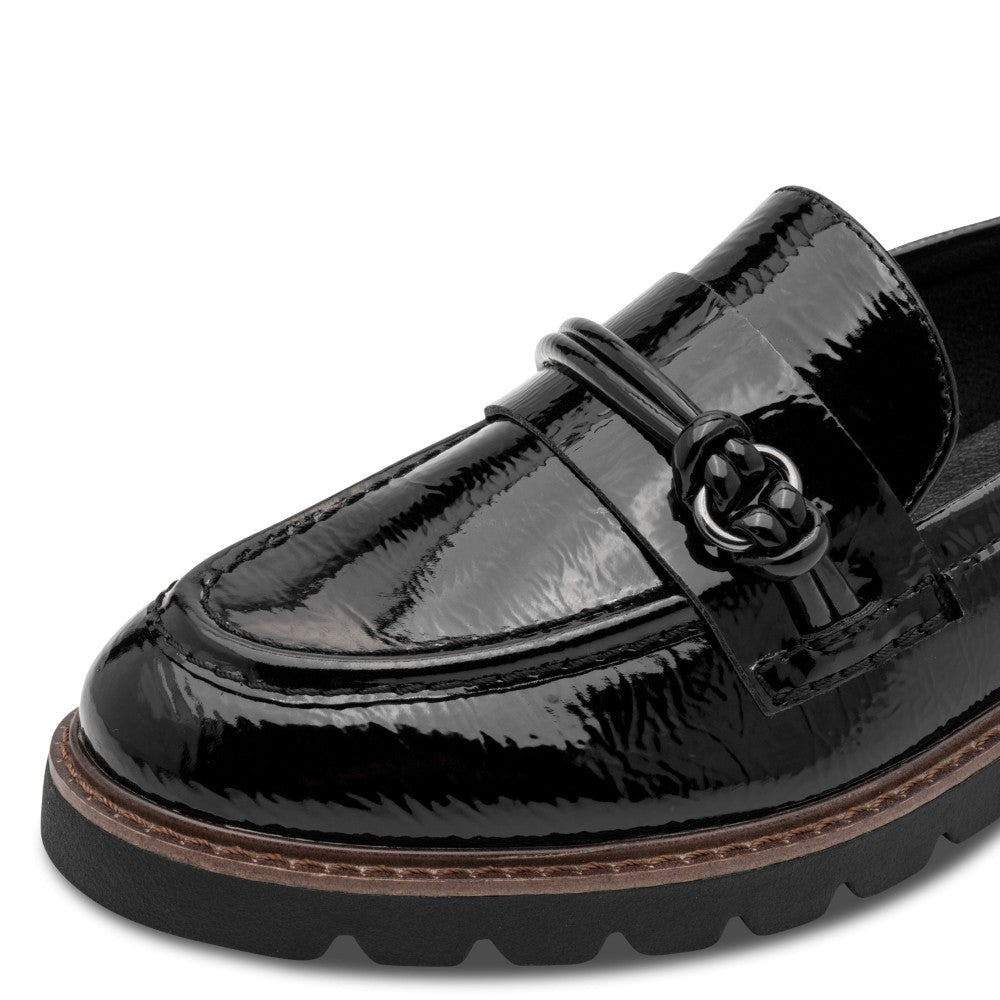 A close-up of a Marco Tozzi black patent leather loafer with a decorative strap and ring detail, a brown welt, and a black lug sole, shown on a white background.