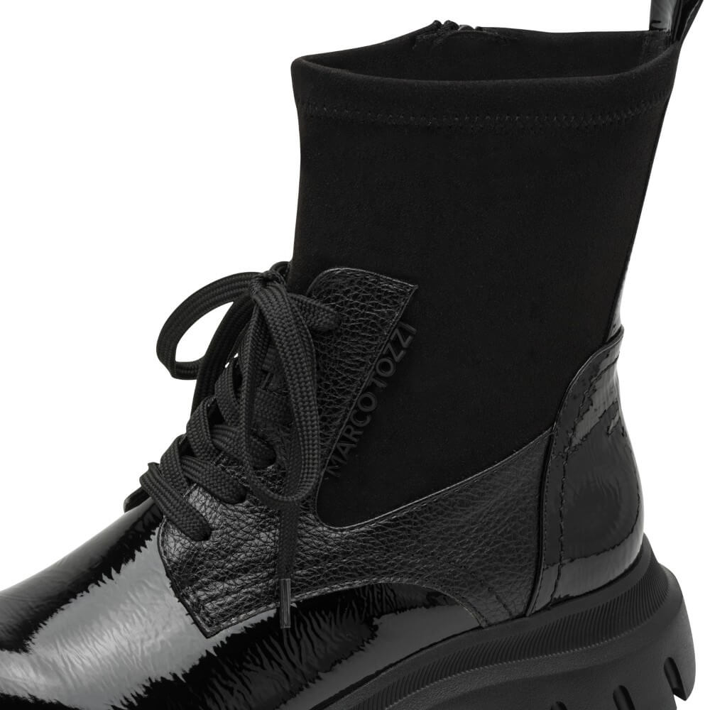 A Marco Tozzi Vegan black ankle boot with a glossy patent leather toe, textured side panel, black laces, a side zipper, and a chunky lug sole, shown against a plain white background.