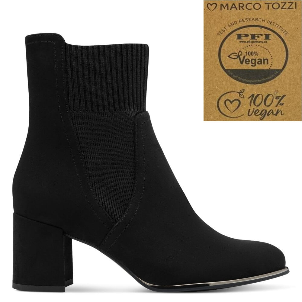 A black ankle boot with a ribbed elastic side panel and a chunky block heel, shown in profile on a white background. A brown cardboard-style badge in the top right reads 'Marco Tozzi' and '100% vegan' with a PFI test logo.