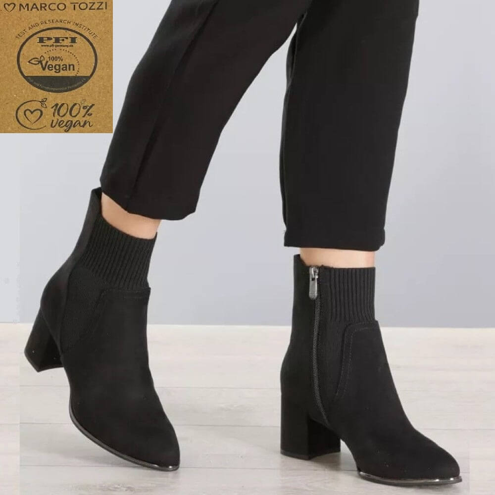 Black ankle boots with a ribbed knit sock-style collar and a block heel, shown on a model wearing black pants. A brown paper badge in the top-left corner reads 'Marco Tozzi' and '100% vegan'.