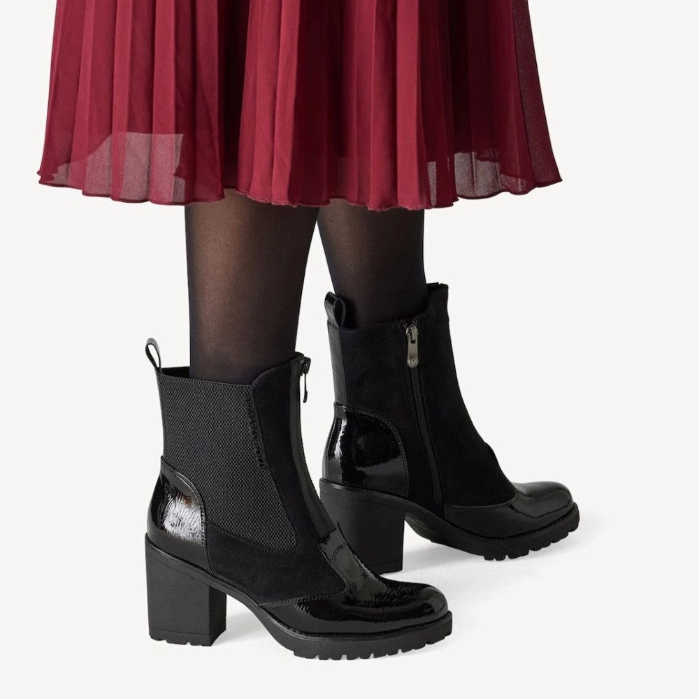 Marco Tozzi Black ankle boots with a block heel, side zipper, and glossy patent leather detail, shown on a model wearing sheer black tights and a pleated red skirt against a light gray background.