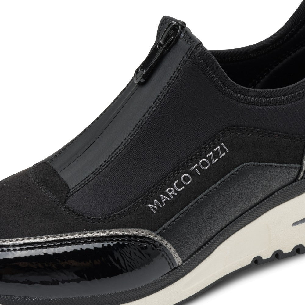 A close-up of a black slip-on sneaker by Marco Tozzi, featuring a side zipper, textured synthetic panels, a glossy patent leather toe cap, and a white midsole.