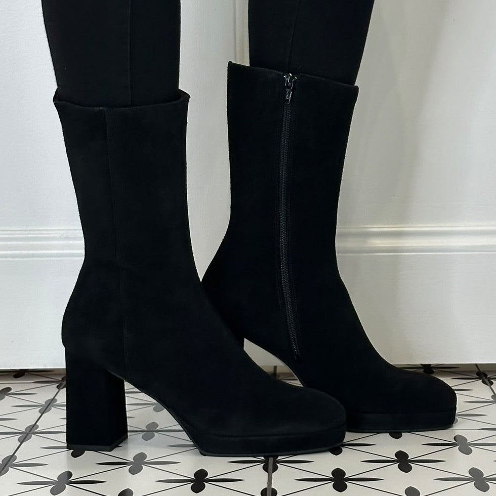 A pair of Marian black suede mid Calf boots with a block heel and side zipper, shown on a person wearing black leggings against a white wall and patterned tile floor.