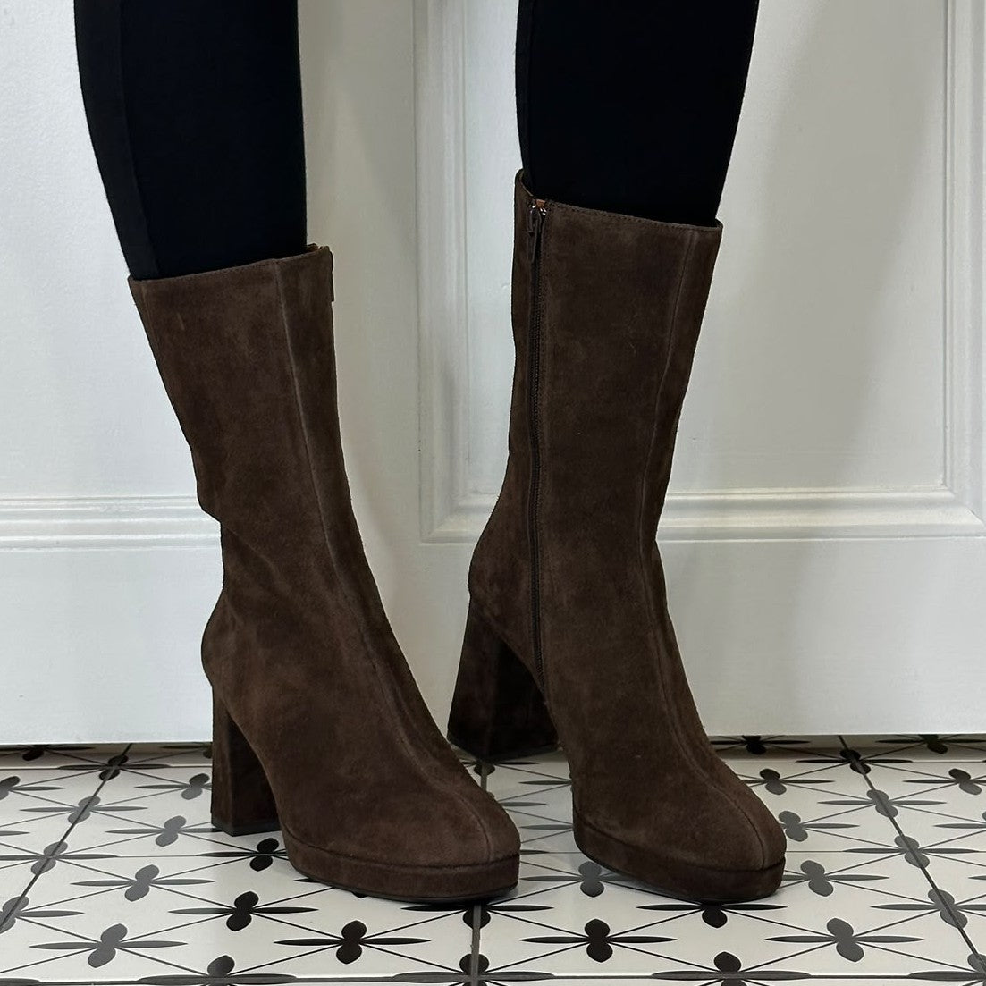 A pair of Marian dark brown suede mid-calf boots with block heels and side zippers, shown on a model wearing black leggings against a white panelled wall and patterned tile floor.