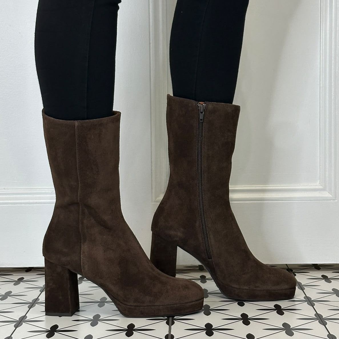 A pair of Marian dark brown suede mid-calf boots with block heels and side zippers, shown on a model wearing black leggings against a white panelled wall and patterned tile floor.