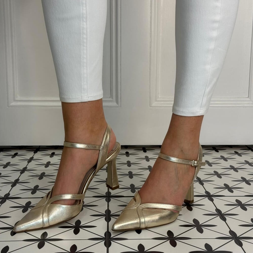 Marian Gold high-heeled shoes worn with white pants on a patterned floor.