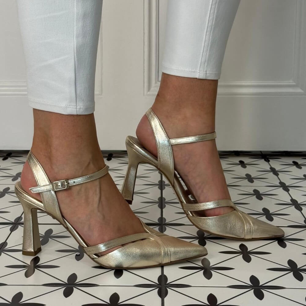 Marian Gold high-heeled shoes worn with white pants on a patterned floor.
